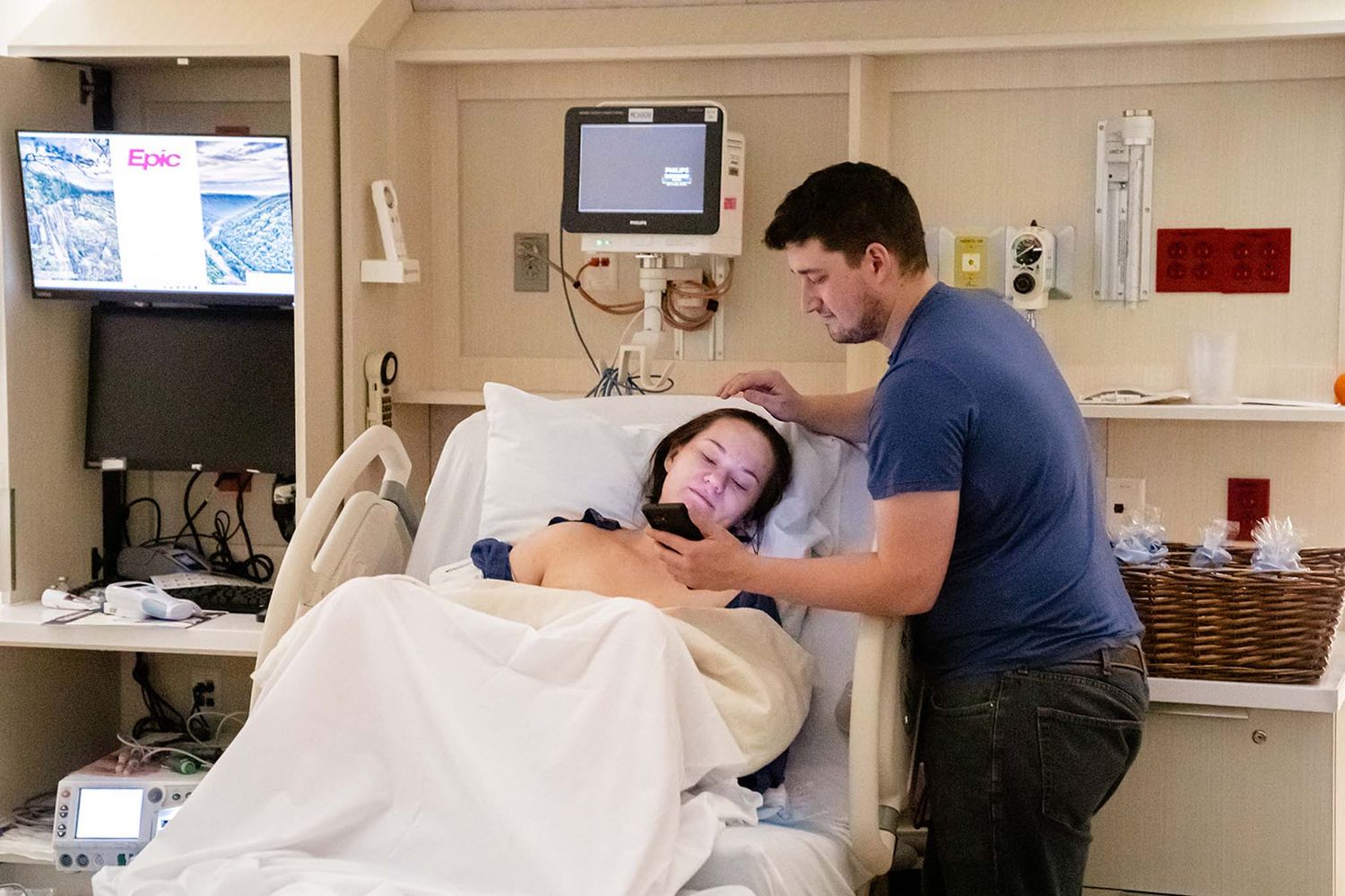 A man tenderly comforts a woman lying in a hospital bed, gently stroking her hair in a medical room.