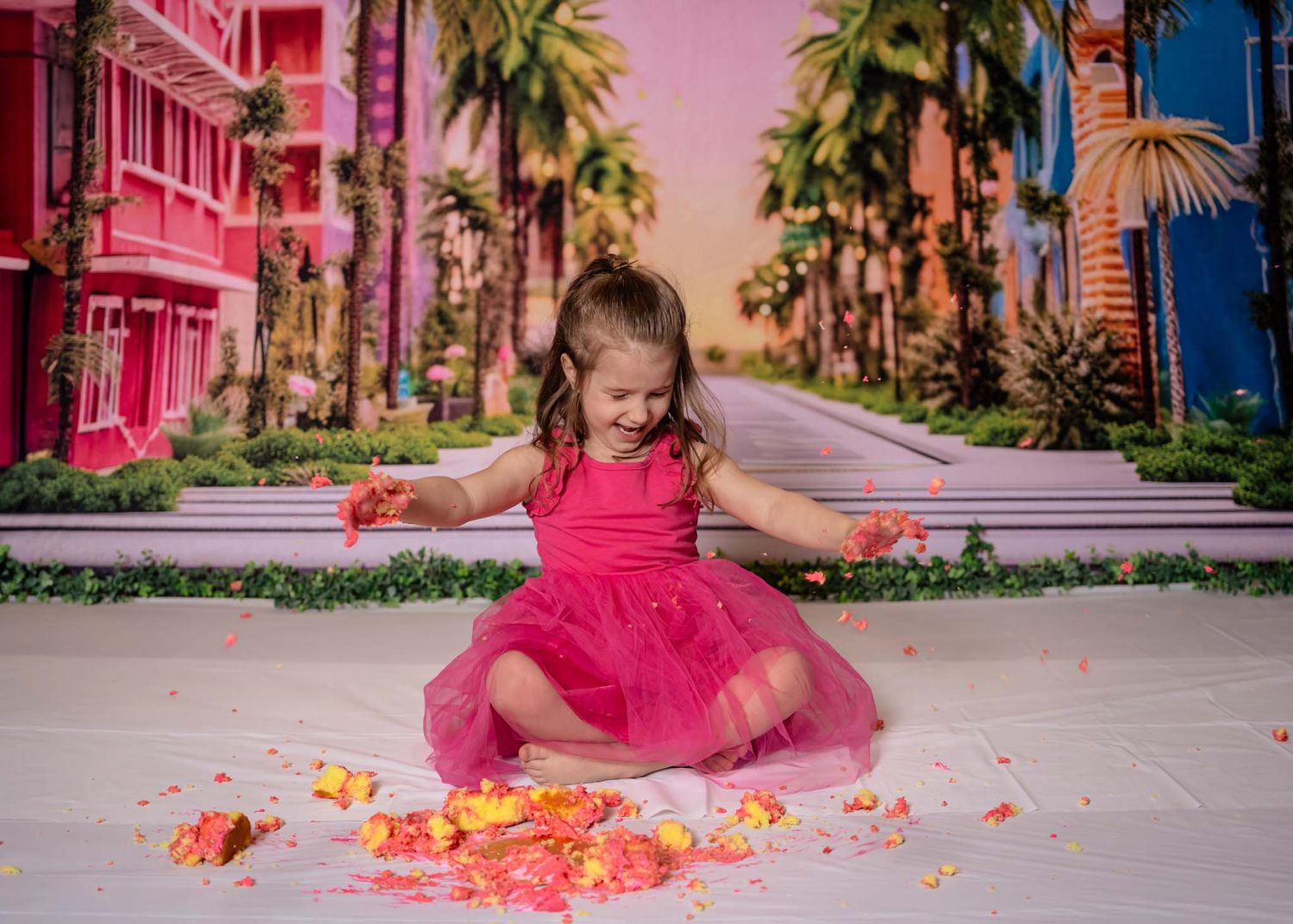 A toddler girl in a pink tutu dress smashes cake on a white floor with a colorful Barbie-themed backdrop.