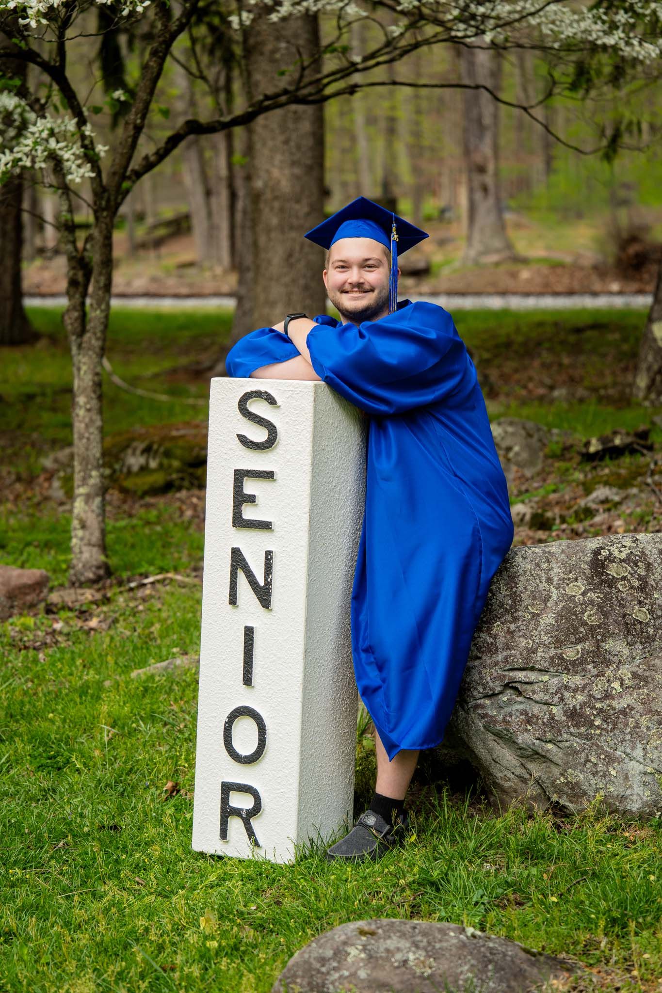 Smiling graduate in blue cap and gown leaning on Senior pillar outdoors in spring.