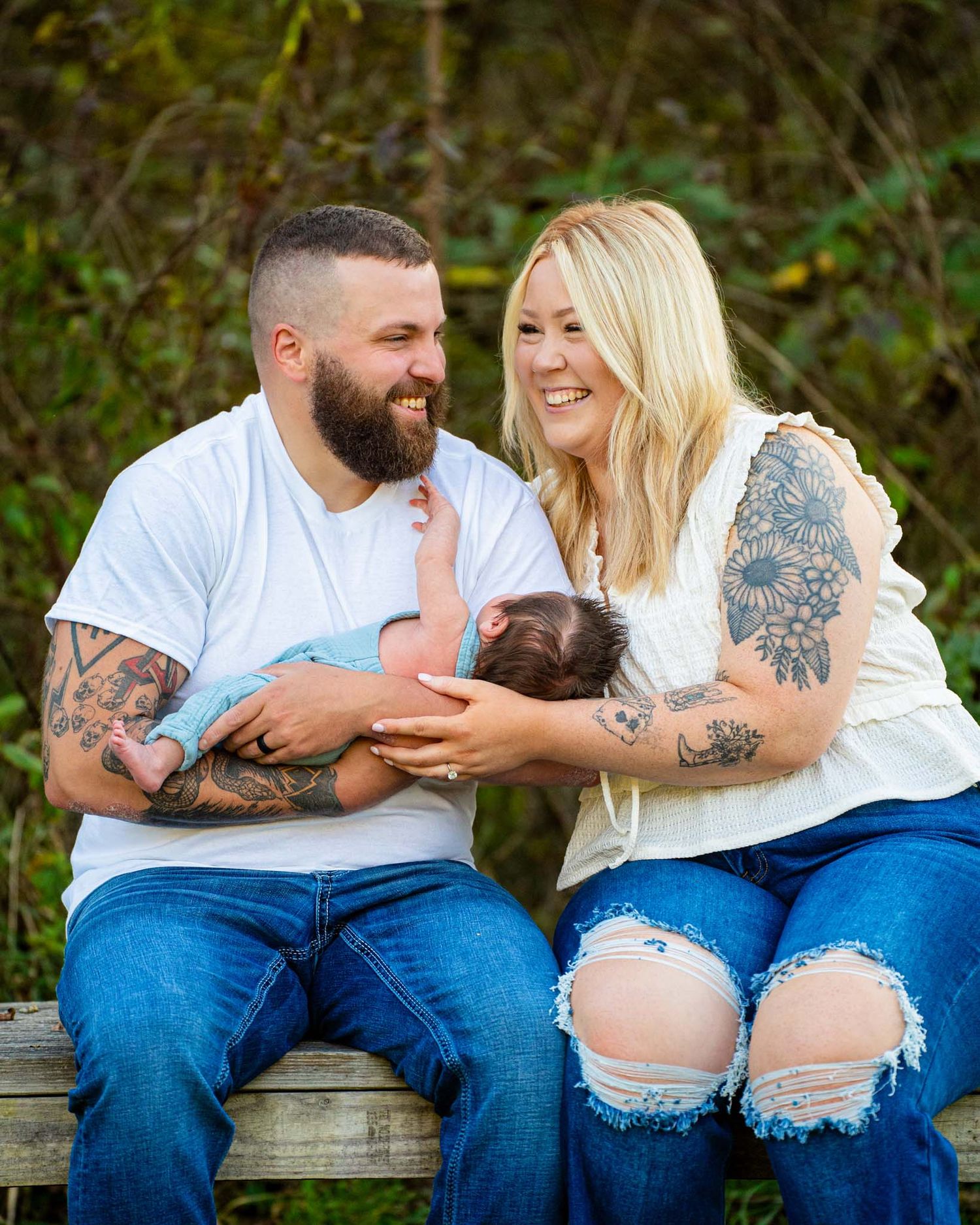 Tattooed couple in white tops and jeans sitting on bench smiling at newborn baby together.