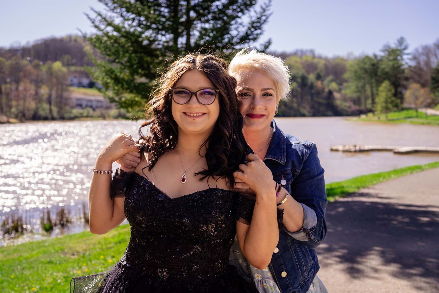 Two women posing together outdoors by a lake, one in a black formal gown and one in denim jacket.