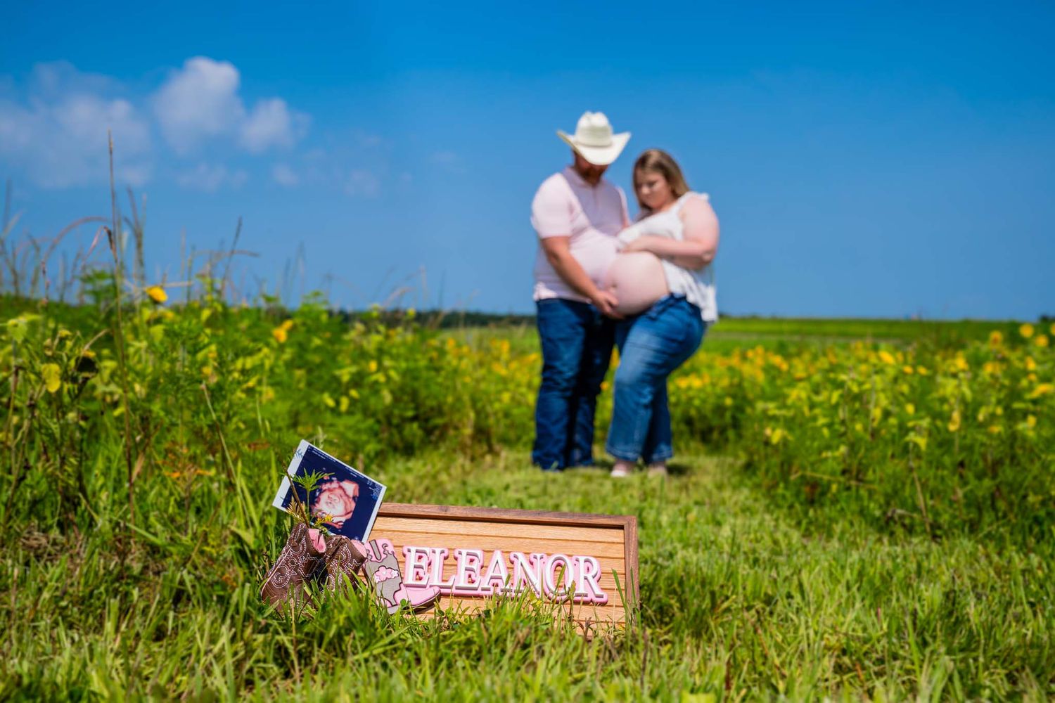 Couple embraces in sunflower field during maternity shoot with Eleanor name sign in foreground.