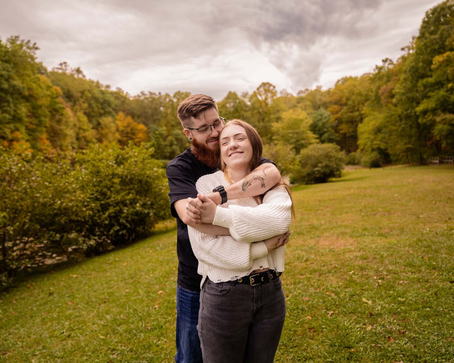 Couple embracing and smiling in a lush green park during autumn, with colorful trees in the background.