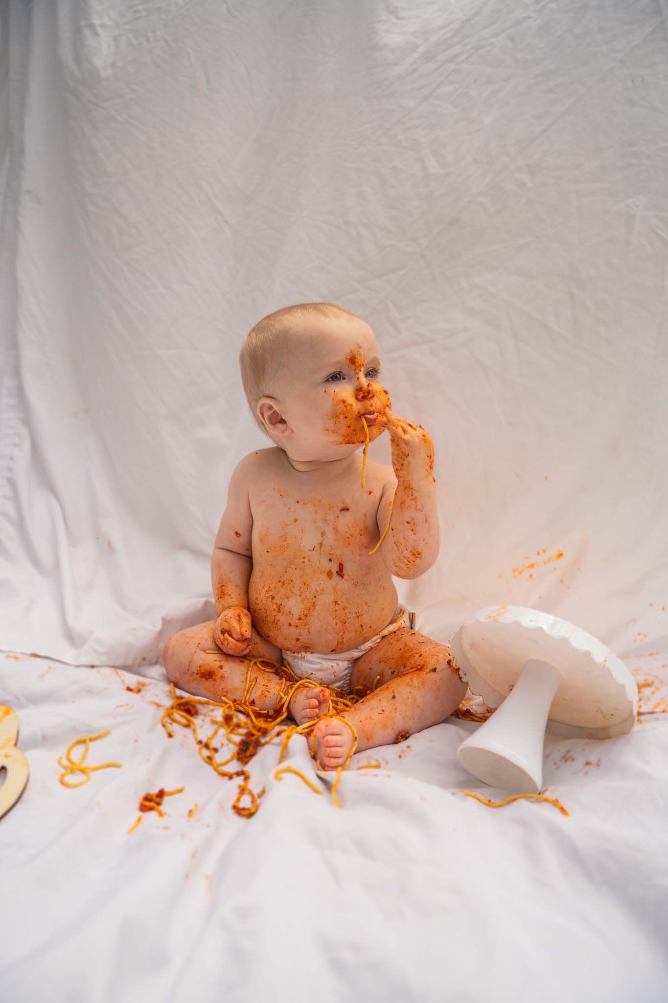 Messy baby covered in orange food sauce sitting on white fabric during a fun first birthday cake smash photo session.