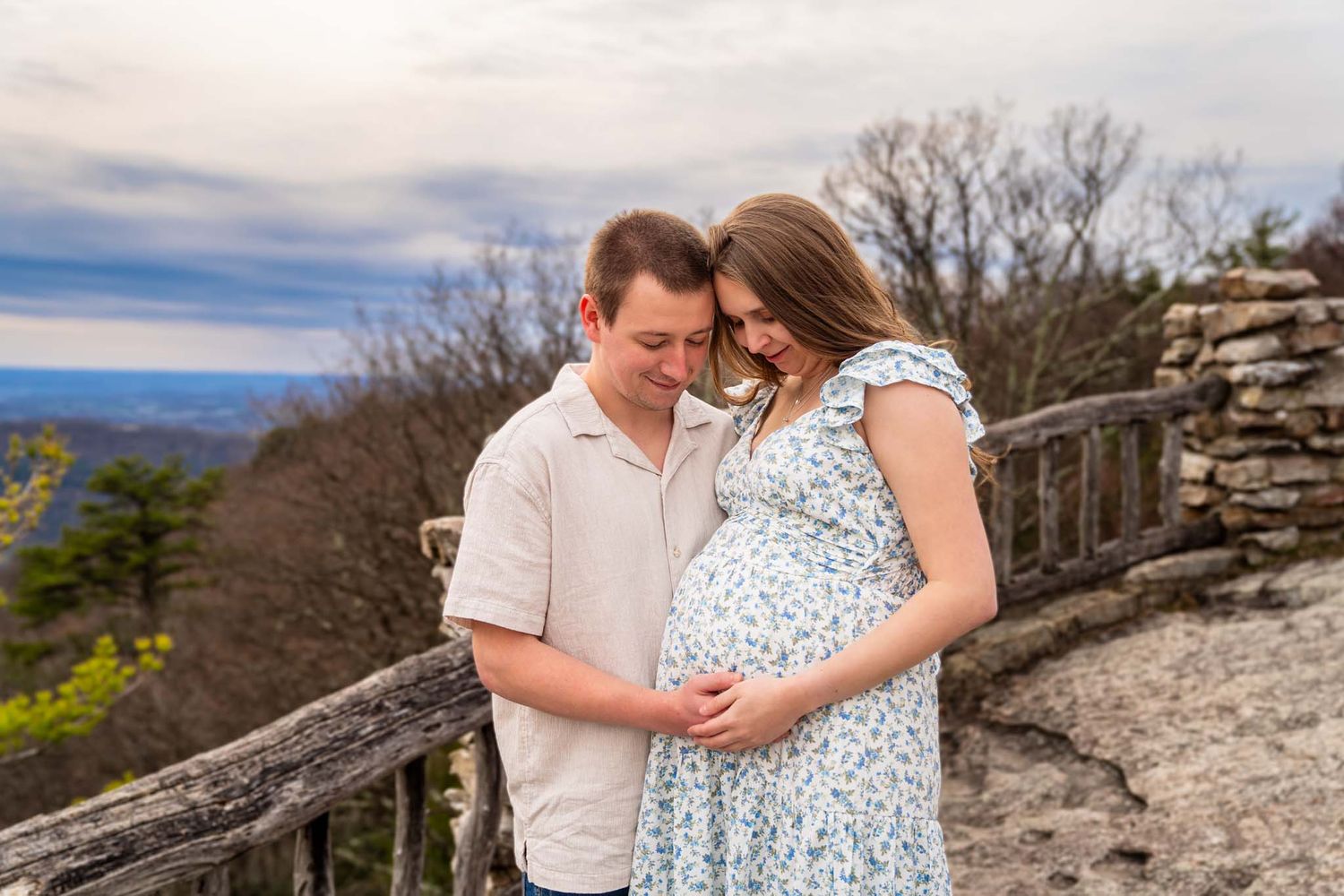 Couple tenderly embracing outdoors on a scenic overlook, woman in floral dress showing pregnant belly, cloudy sky backdrop.