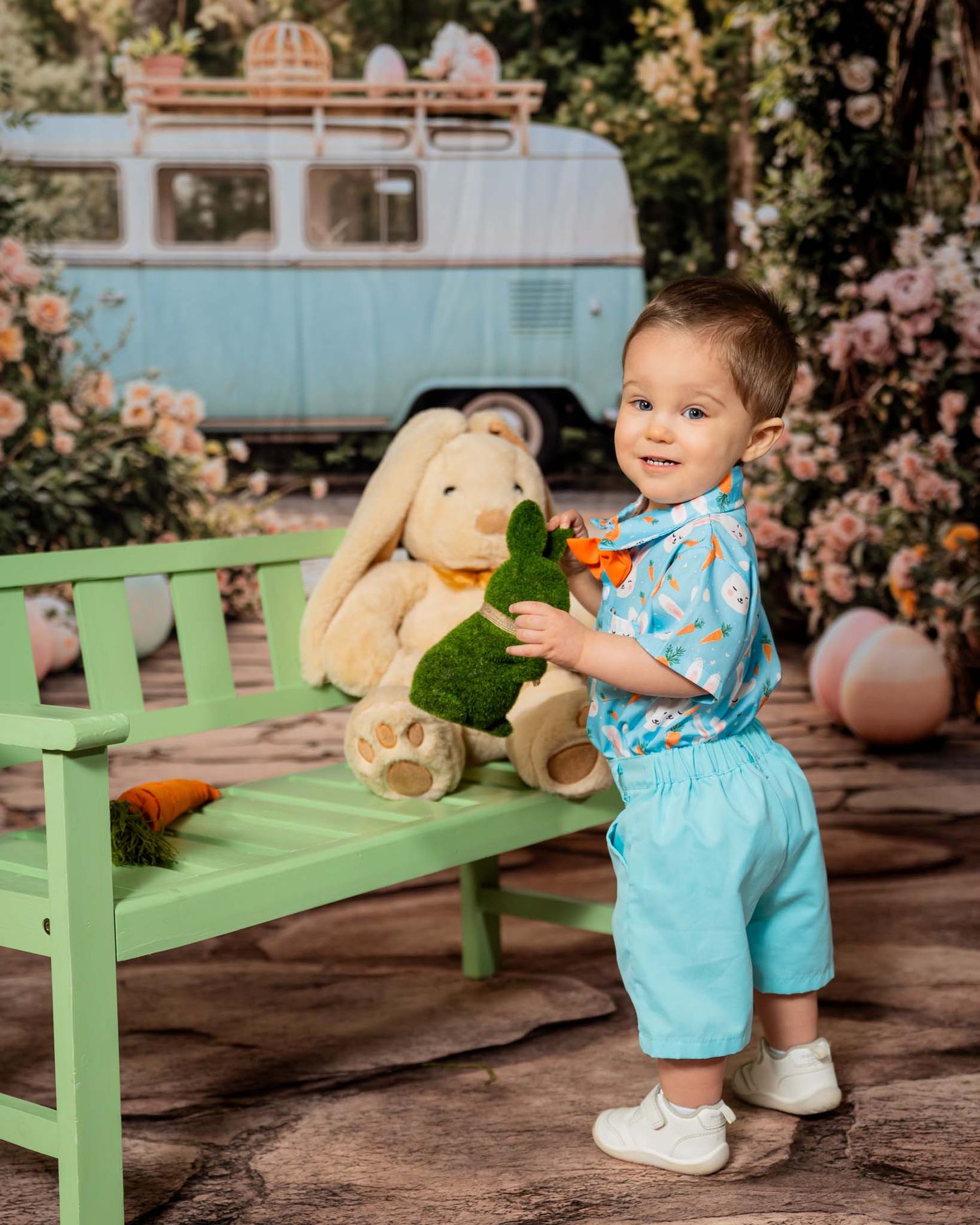 Toddler boy in blue outfit holds green bunny beside a stuffed rabbit on a green bench with a vintage van backdrop.