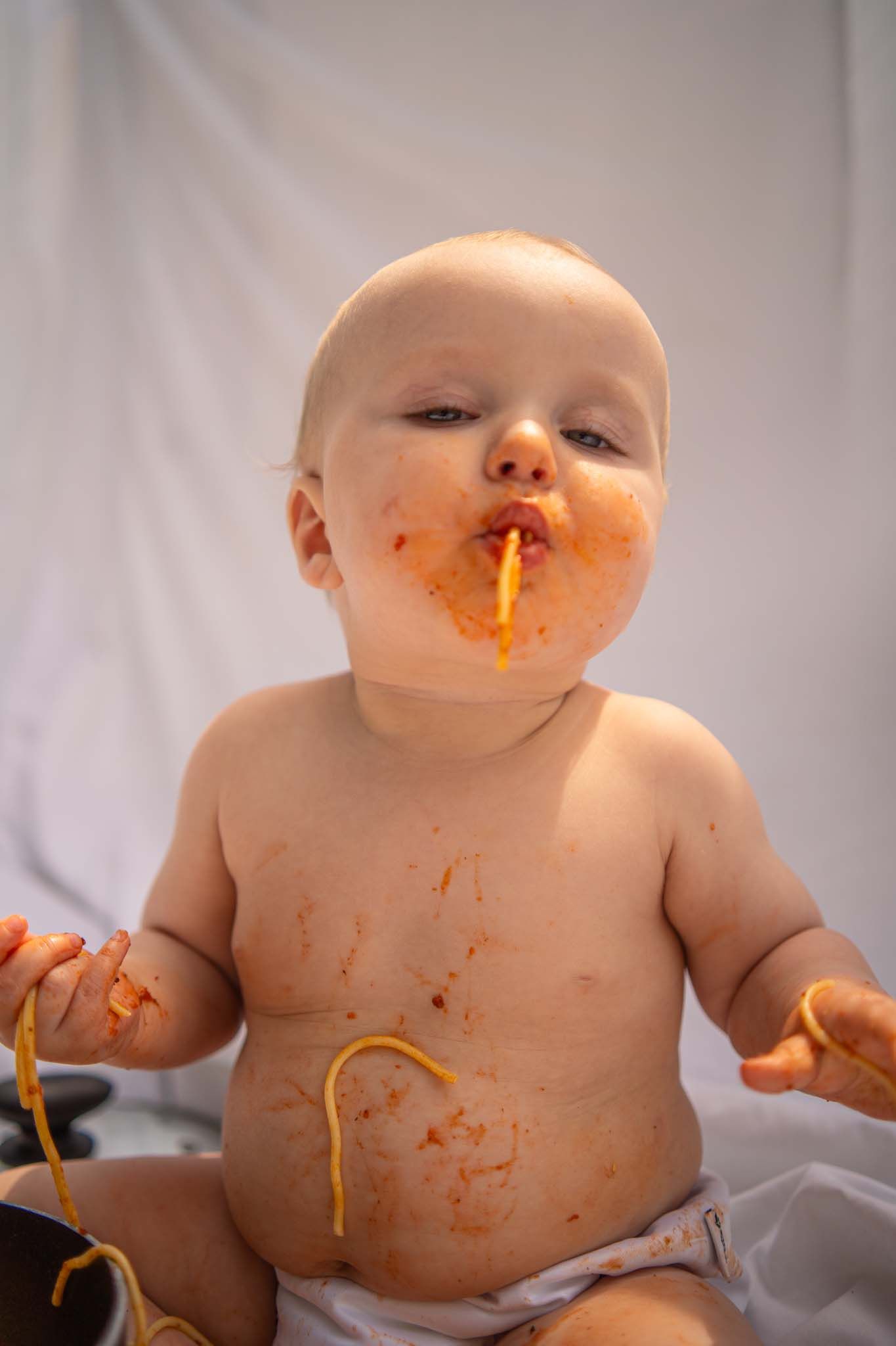 A messy baby covered in spaghetti and tomato sauce, holding noodles while eating with a happy expression.
