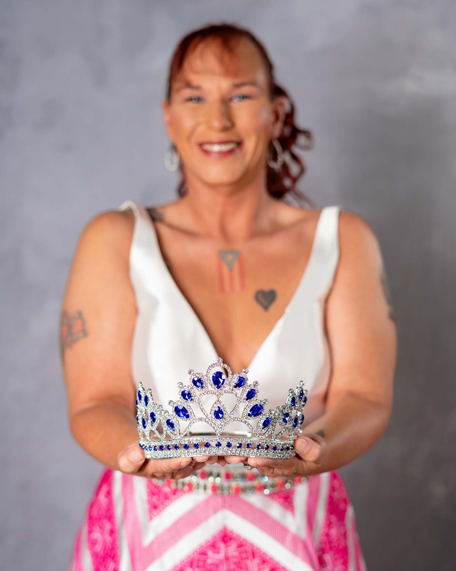 Smiling woman in white dress holding blue gemstone tiara crown toward camera.