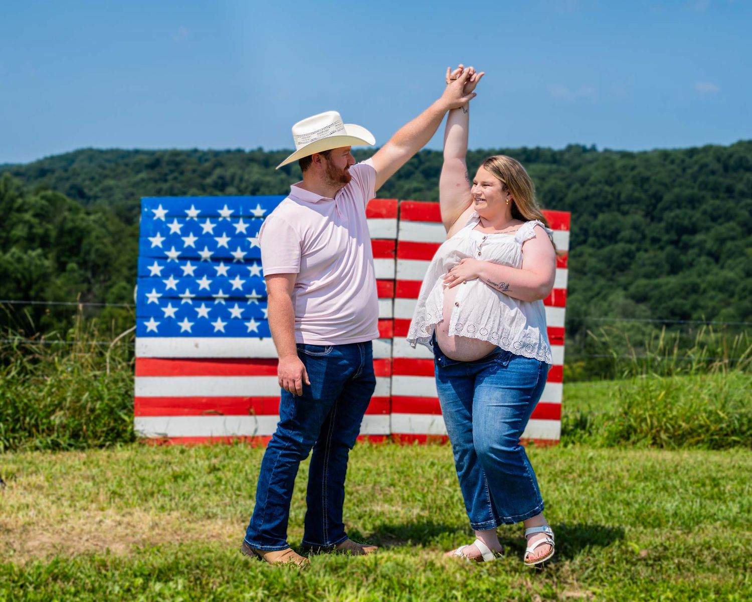 A couple dances together in front of a large rustic American flag on a sunny day.