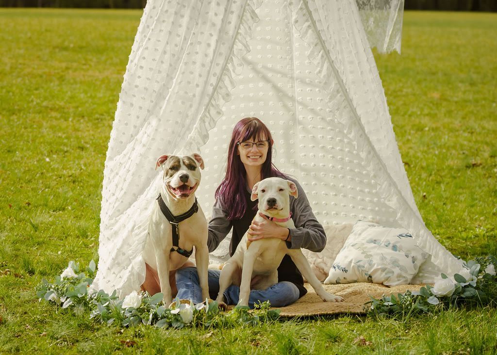 Woman with purple hair sits with two white dogs inside a lace teepee on grass.