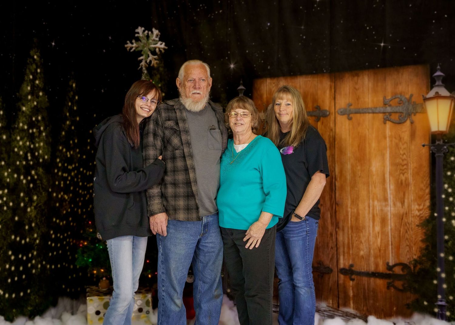 Family of four posing together in front of a rustic wooden door with Christmas tree decorations in the background.