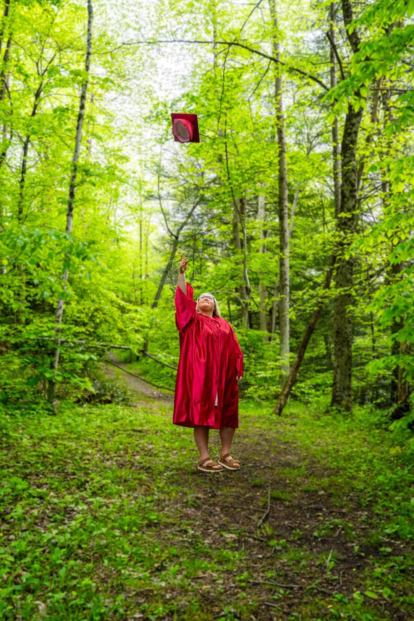 Graduate in red gown tosses cap into air on lush forest trail.