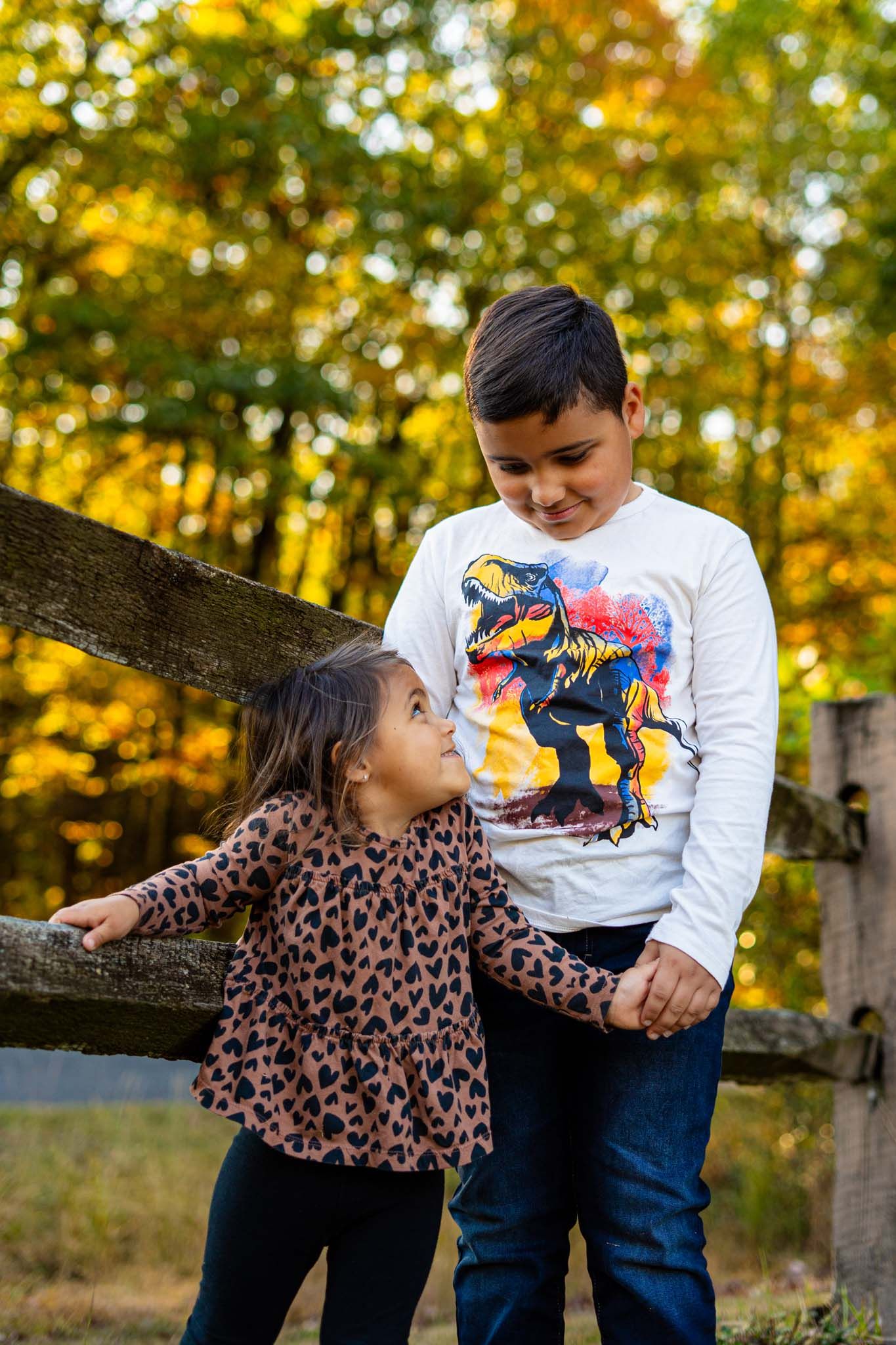 Older brother and younger sister hold hands by wooden fence surrounded by colorful autumn foliage.