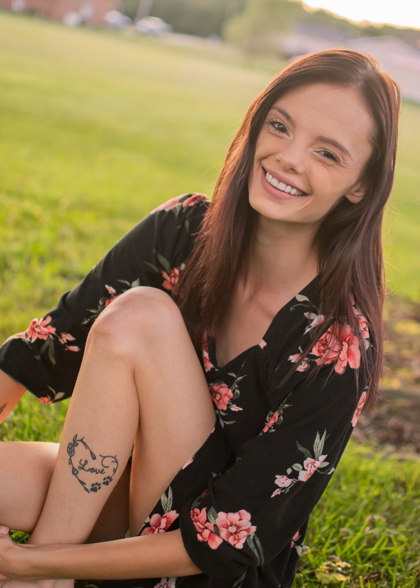 Smiling young woman in floral dress sitting on grass with a small tattoo visible.