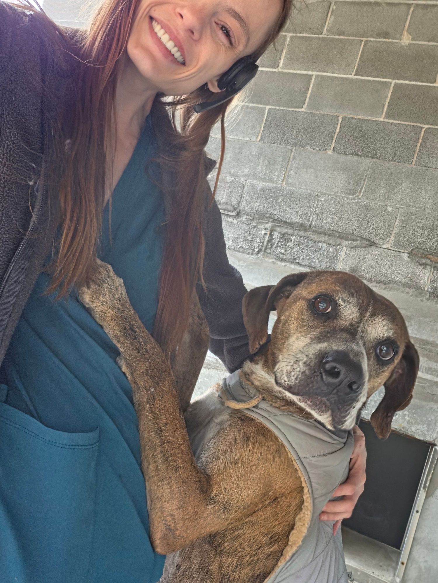 Smiling woman in teal scrubs holds a brown brindle beagle mix dog while taking a selfie outdoors.