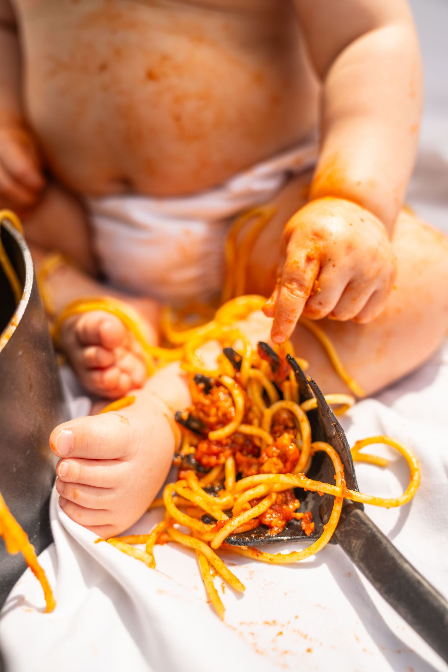 Messy baby playing with spaghetti in tomato sauce, hands and feet covered in orange pasta.