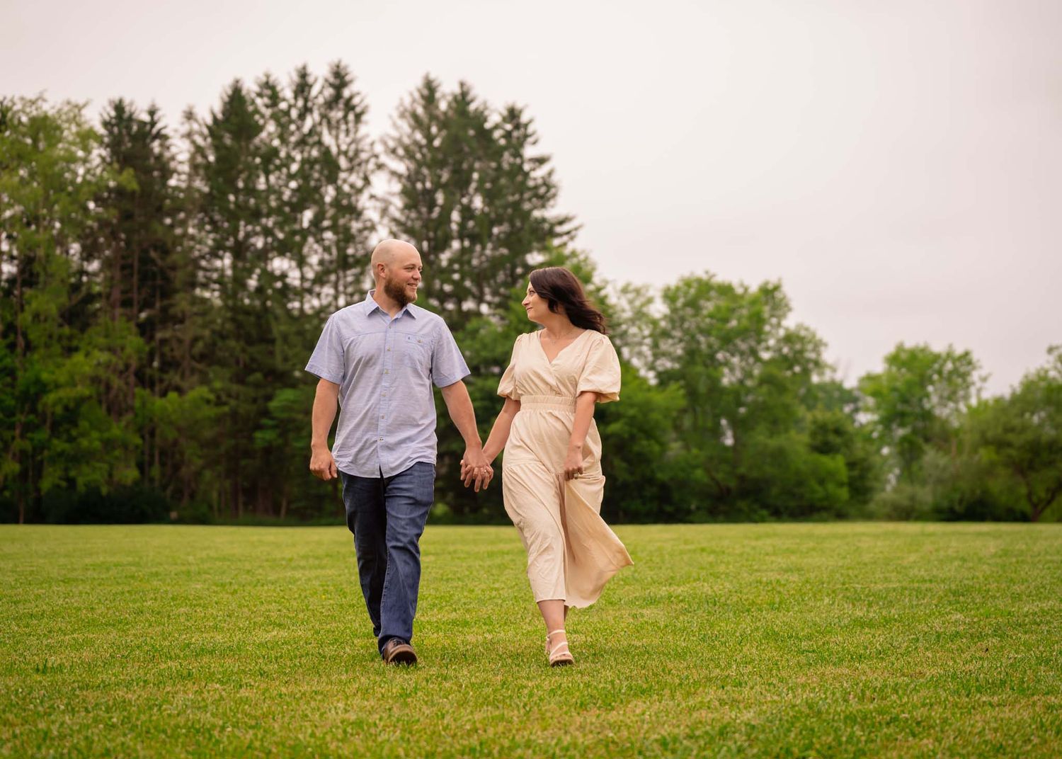 Couple holding hands walking through green grassy field with trees behind.