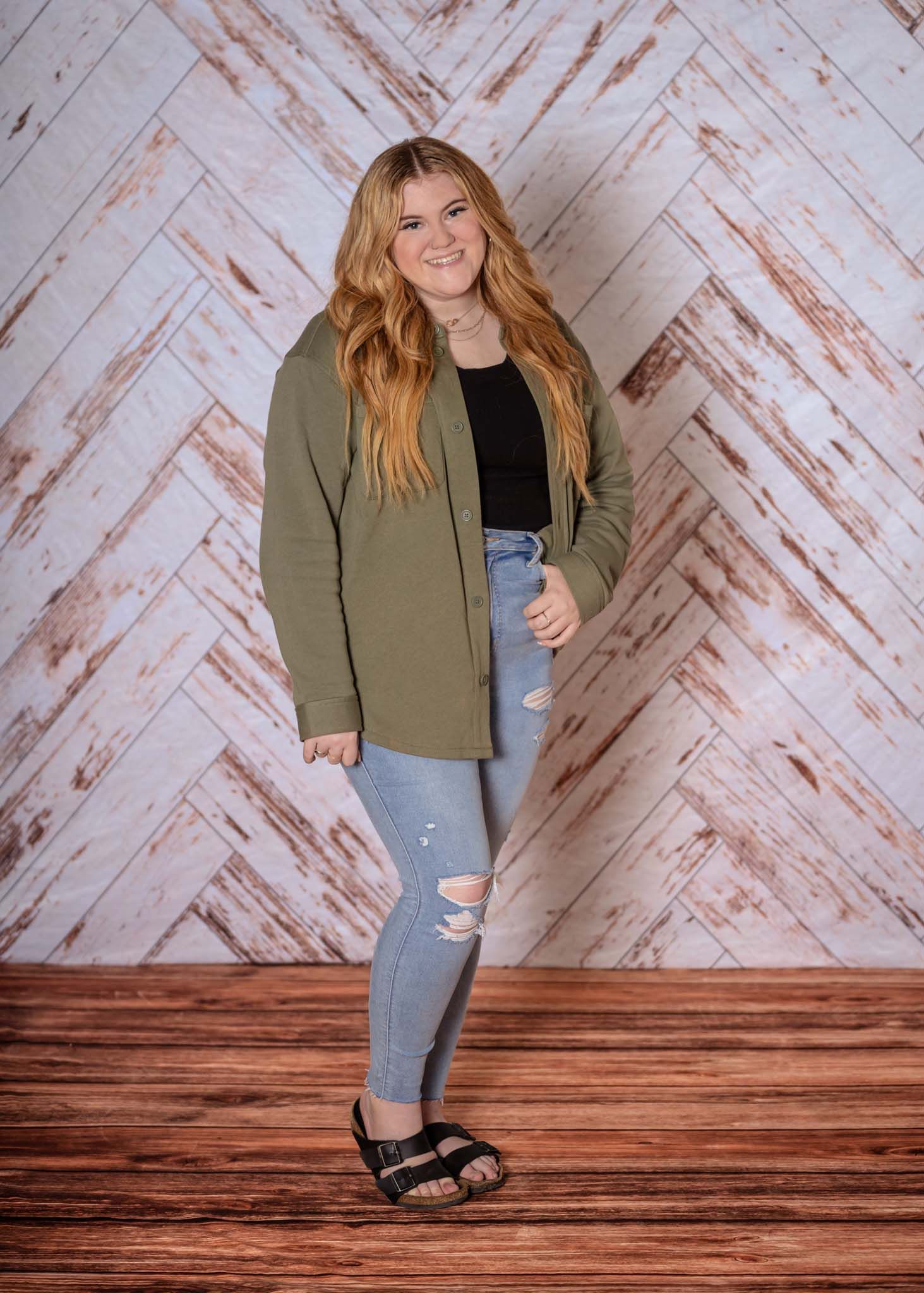 Smiling teen girl with auburn hair in olive blazer and ripped jeans, wooden backdrop.