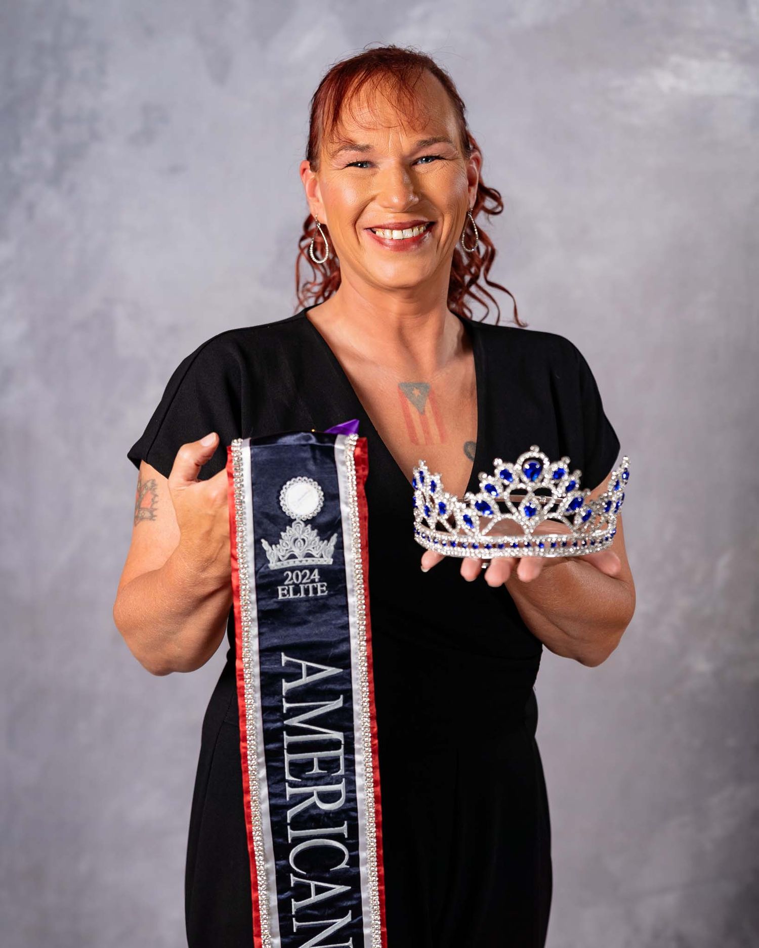 Woman proudly displaying 2024 Elite American Women pageant sash and jeweled crown in studio portrait.