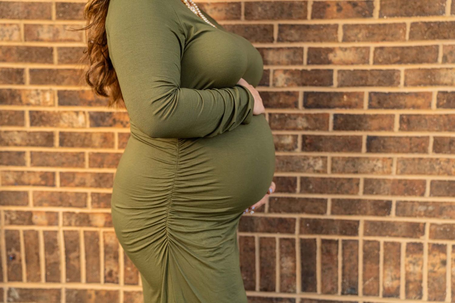 A pregnant woman in an olive ruched dress cradles her baby bump against a brick wall.
