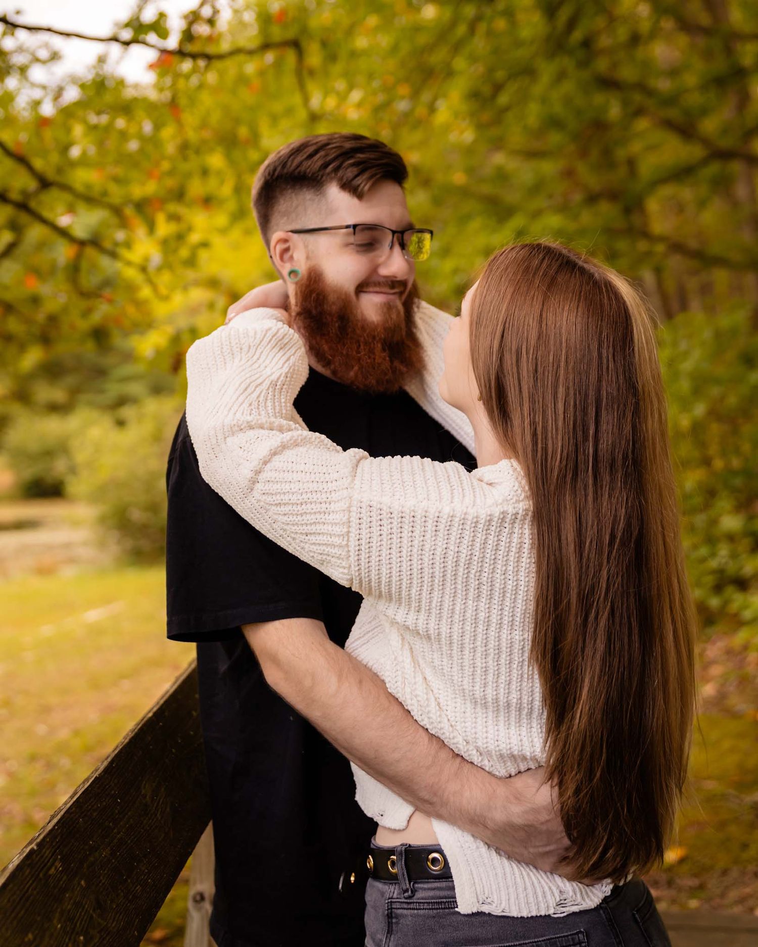 A bearded man with glasses smiles at a woman with long auburn hair as they embrace near a wooden fence outdoors.