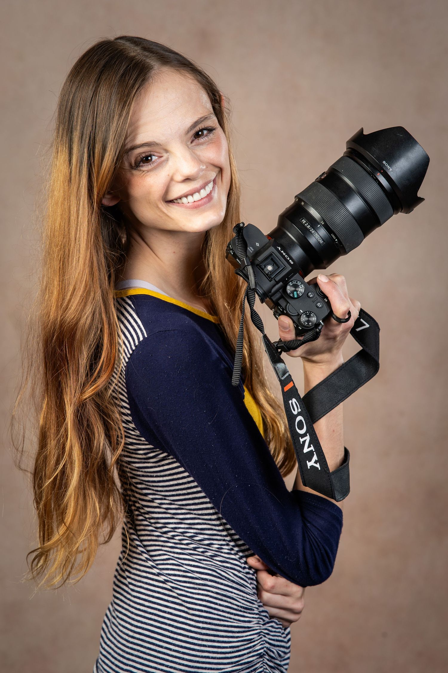 Smiling young woman with long hair holding a Sony camera with lens, wearing a striped shirt in a studio setting.
