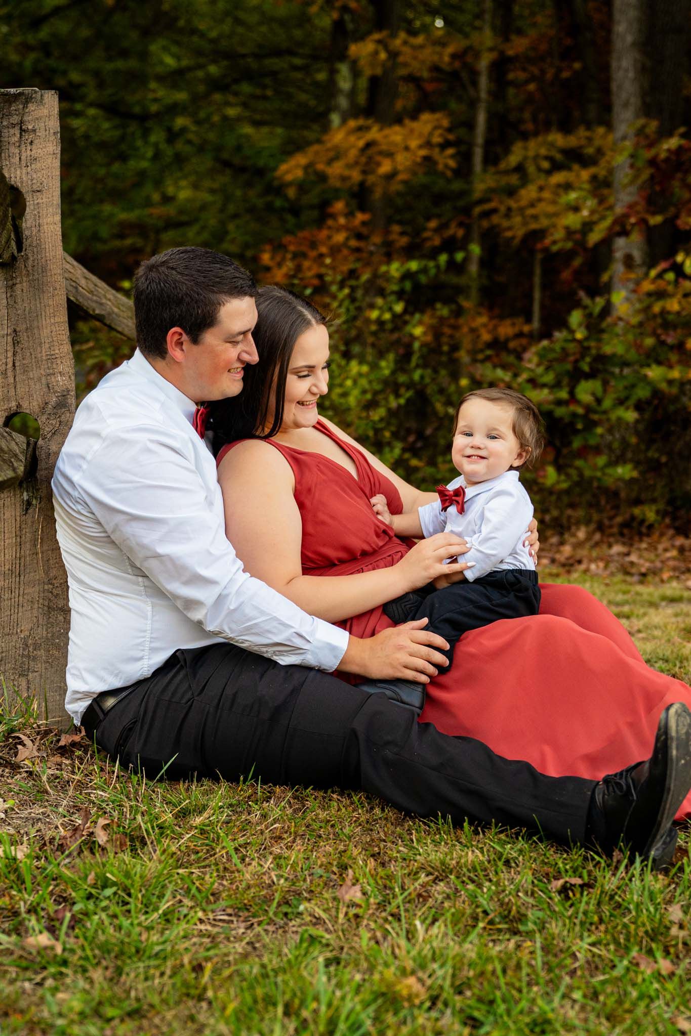 Happy family with baby boy in matching red and white outfits sitting on grass by wooden fence.