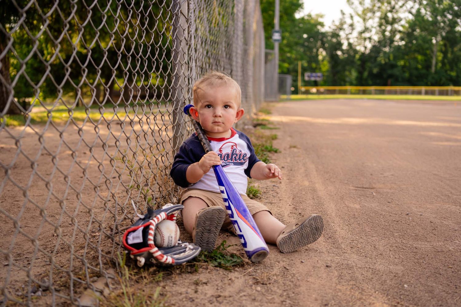 Toddler in baseball uniform sitting by chain-link fence with bat, glove, and ball on dirt field.