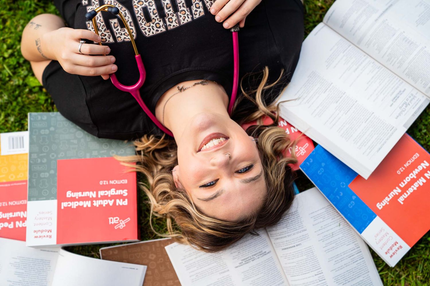Nursing student lying on grass surrounded by medical textbooks with stethoscope.