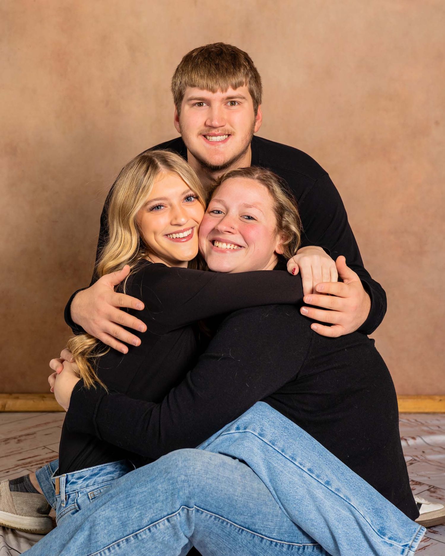 Three siblings hugging and smiling warmly in a studio portrait wearing matching black tops and jeans.
