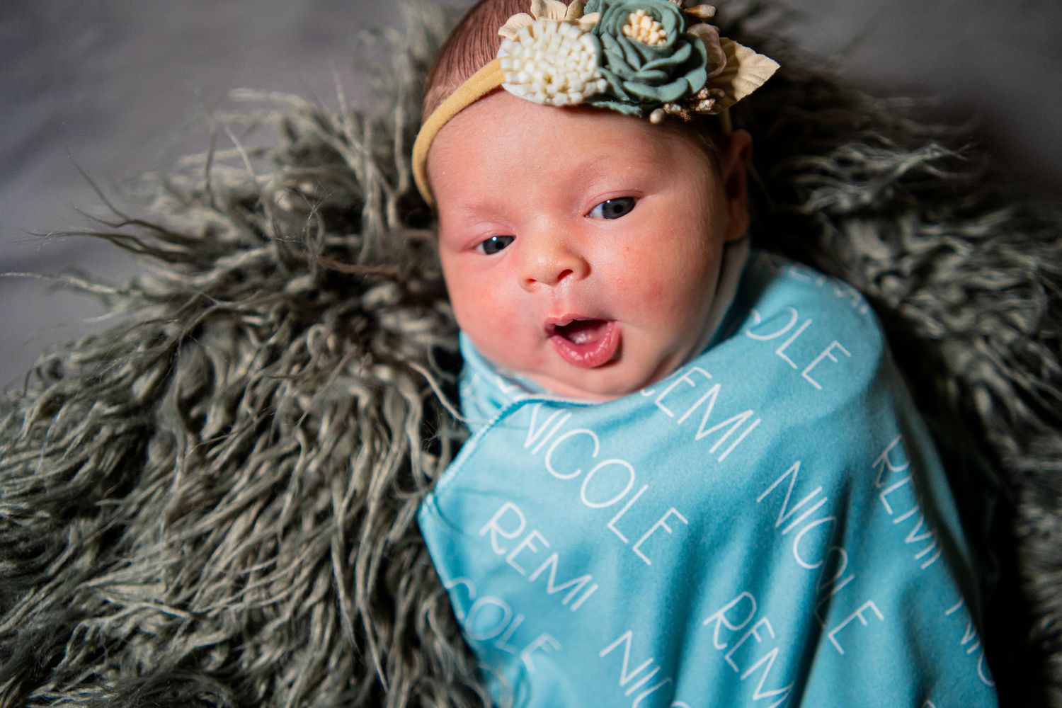 Newborn baby girl swaddled in teal personalized blanket with name Remi Nicole, wearing floral headband.