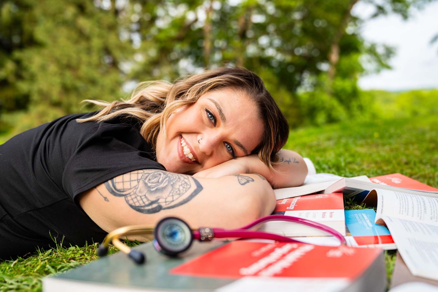 Smiling woman with tattoo lying on grass in park surrounded by medical books and stethoscope.