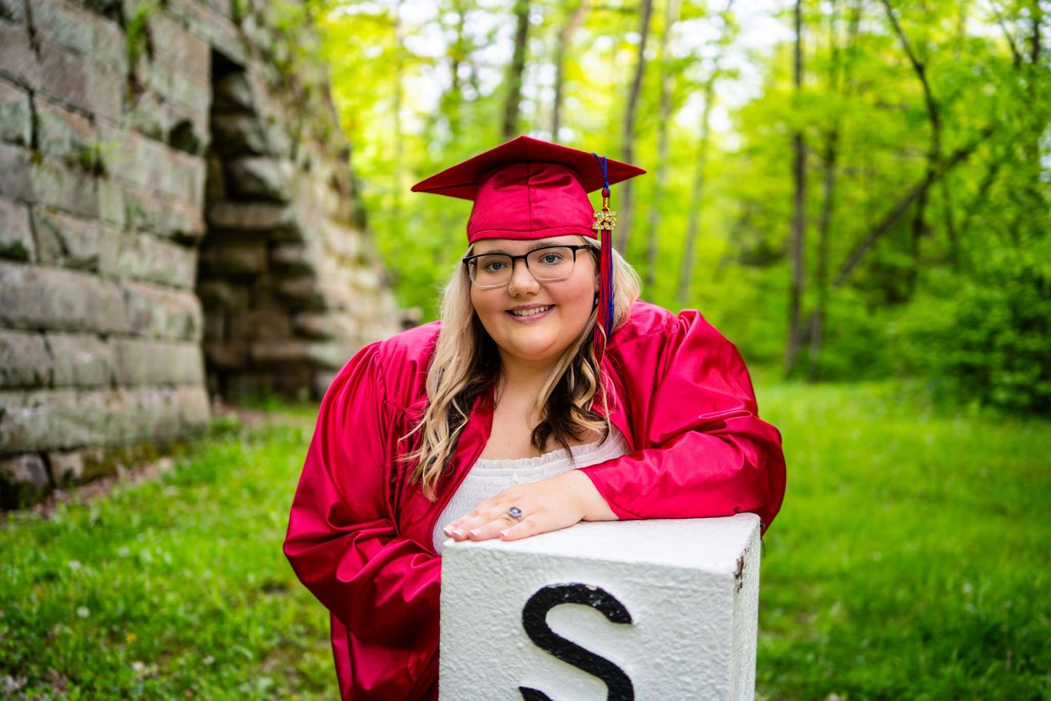 Young woman in red graduation cap and gown posing outdoors with a decorative letter S block in a green park setting.