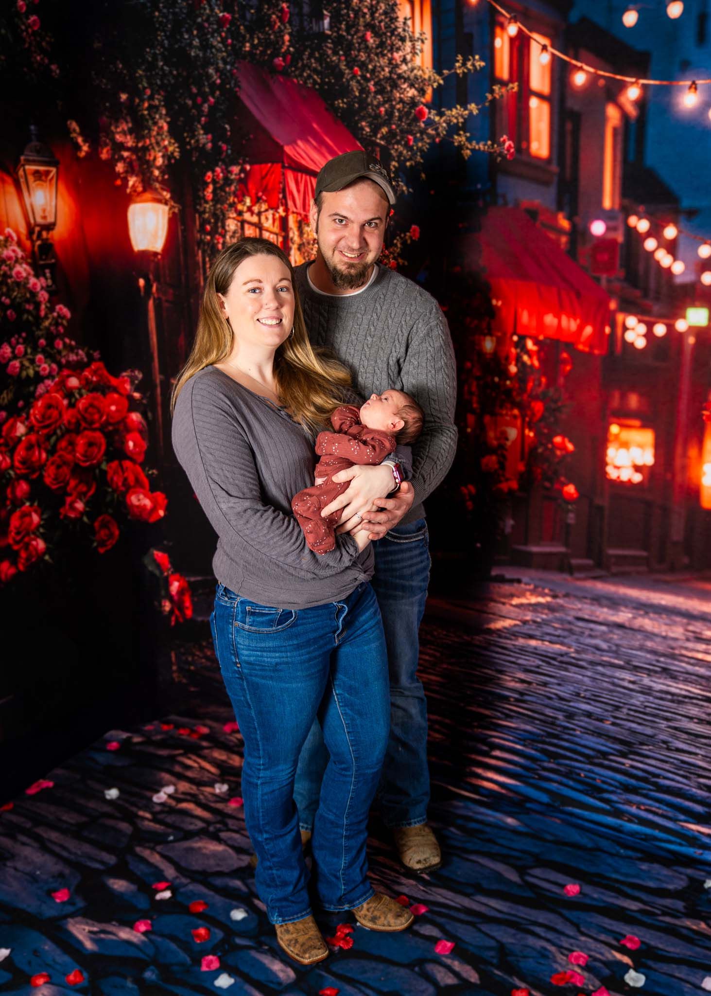 Smiling parents holding their newborn baby on a cobblestone street with red roses at night.