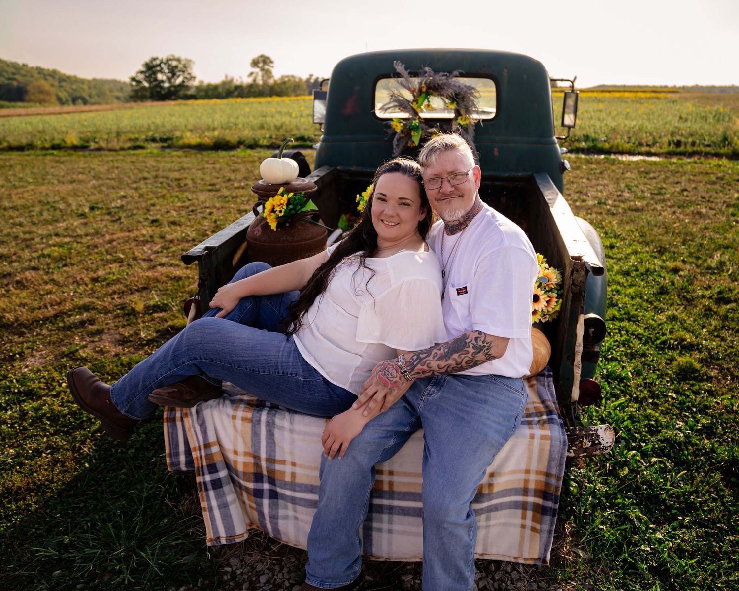 Couple sitting in vintage truck bed surrounded by sunflowers in a green field at sunset.