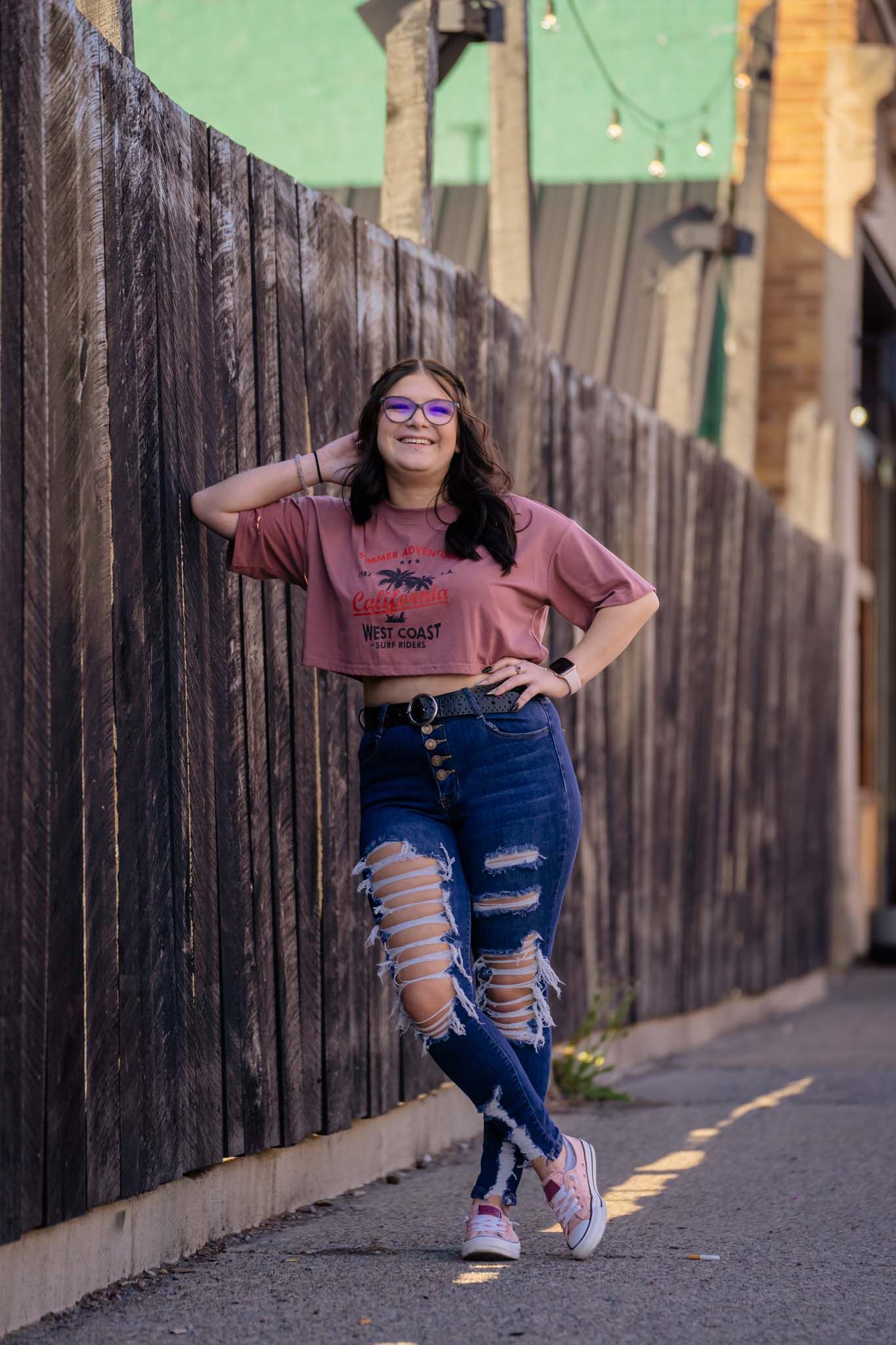 Teen girl in ripped jeans and pink top leaning against a wooden fence in a sunlit alleyway.