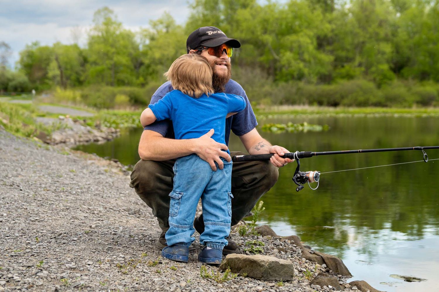 Bearded father hugging young son while holding fishing rod near calm lake shoreline.