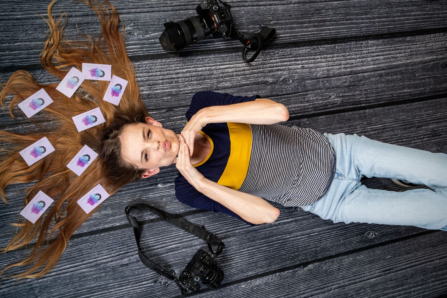 A young woman with long hair lies on wooden floor surrounded by photos and cameras in a creative photography concept.