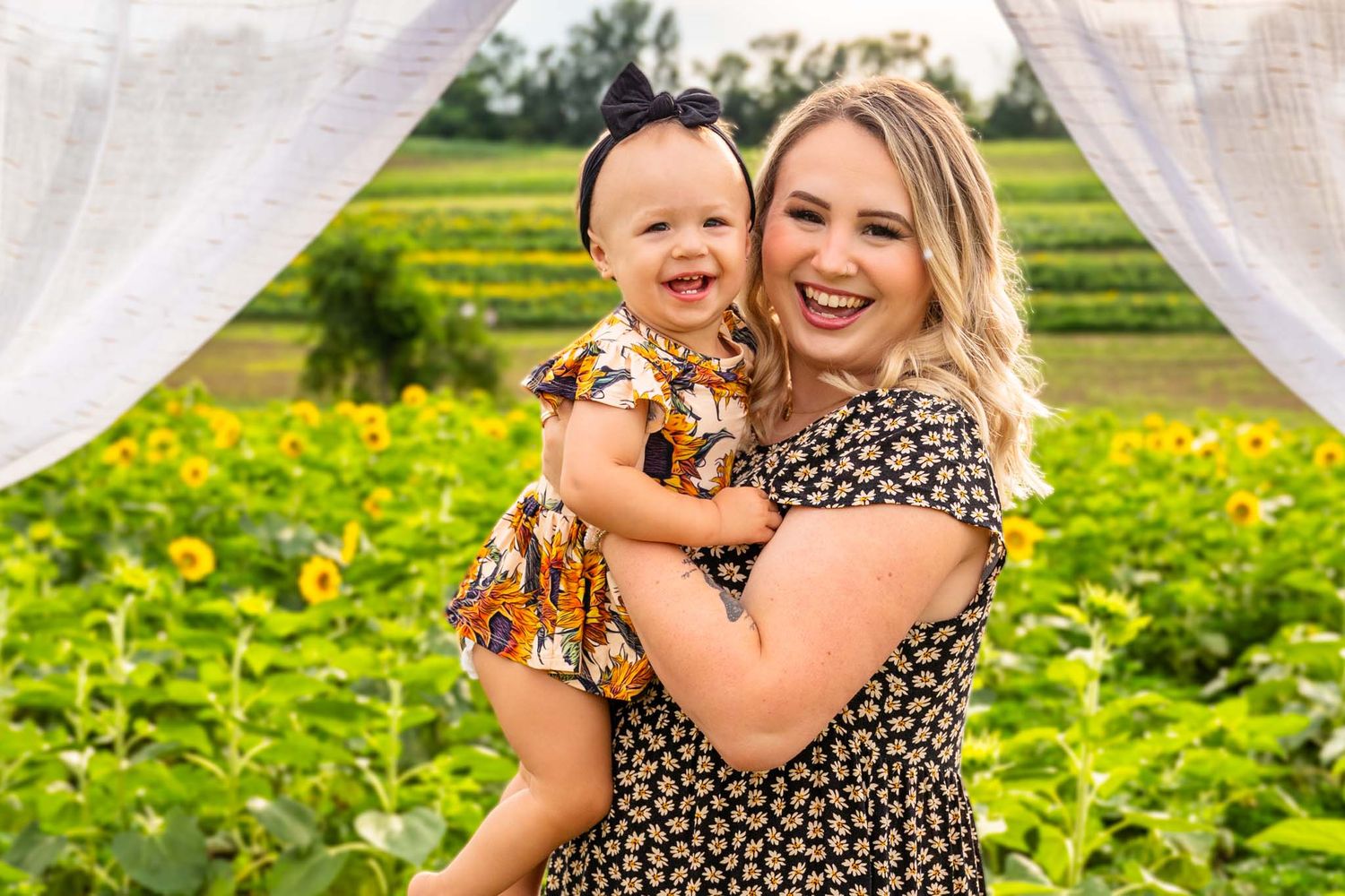 Blonde mother holding laughing toddler girl in sunflower field.