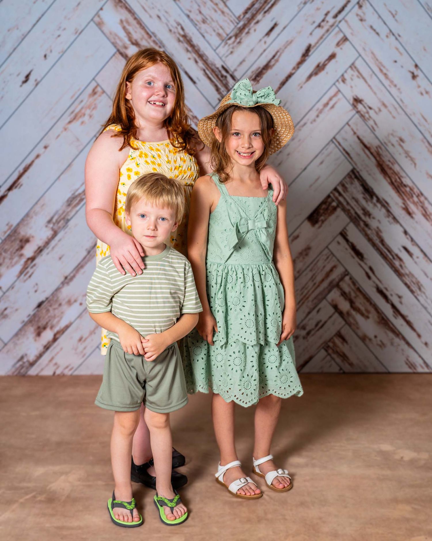 Three children pose for a portrait, wearing coordinating green and yellow spring outfits against a rustic white wood backdrop.