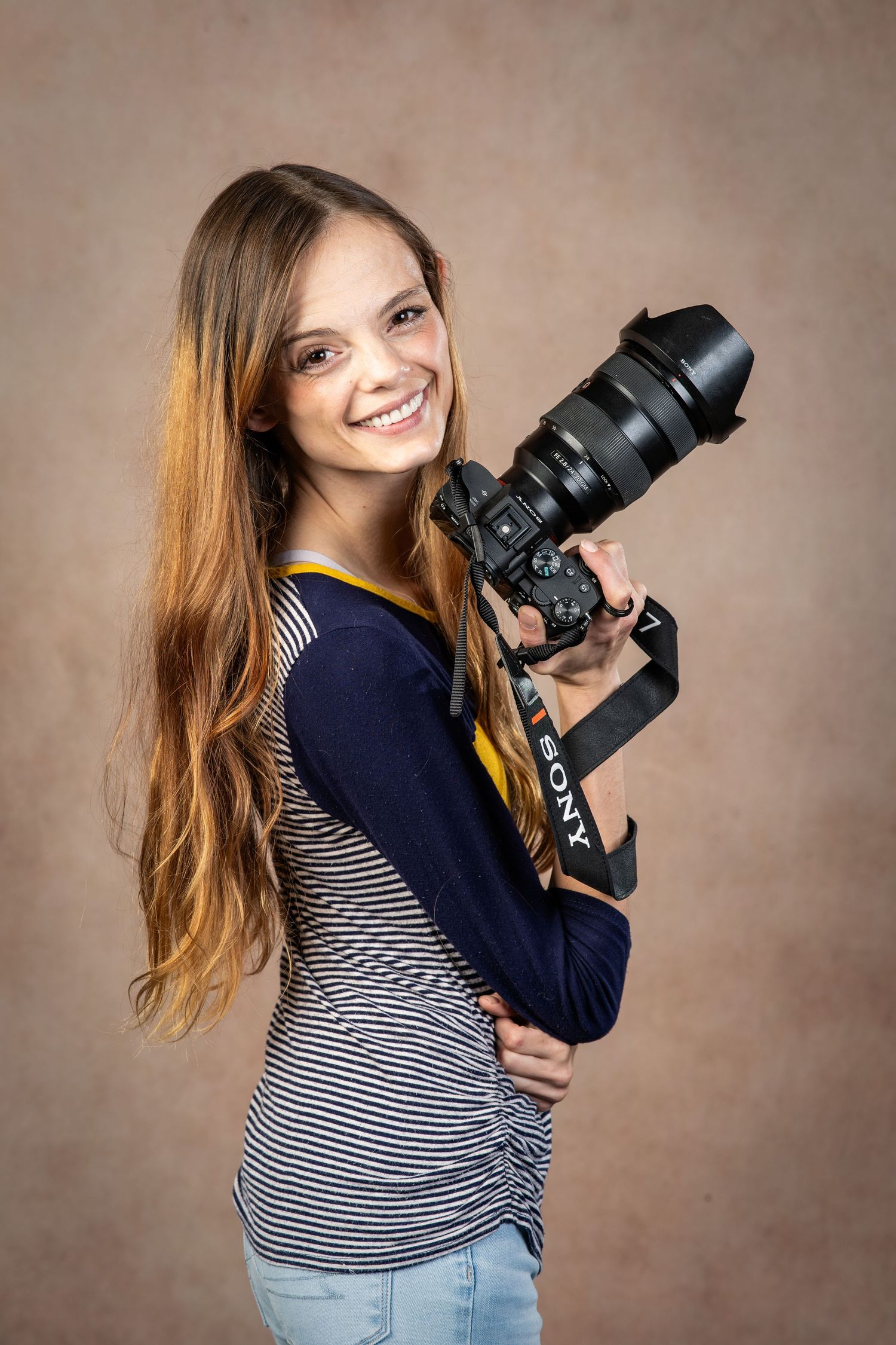 Smiling young woman with long hair holding a Sony DSLR camera with telephoto lens in a studio setting.
