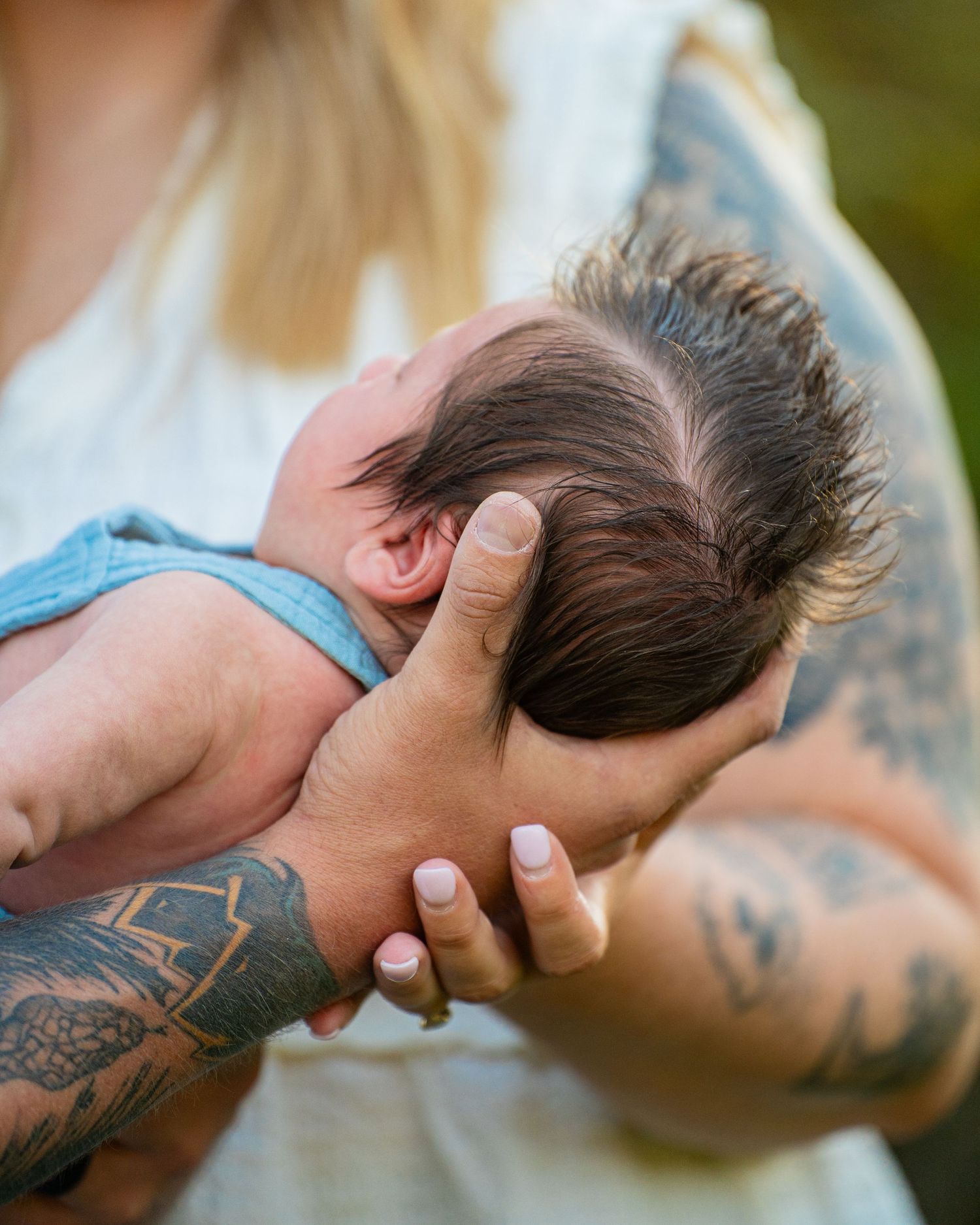 Tattooed arms cradling a newborn baby with dark hair, dressed in a blue onesie, outdoors.