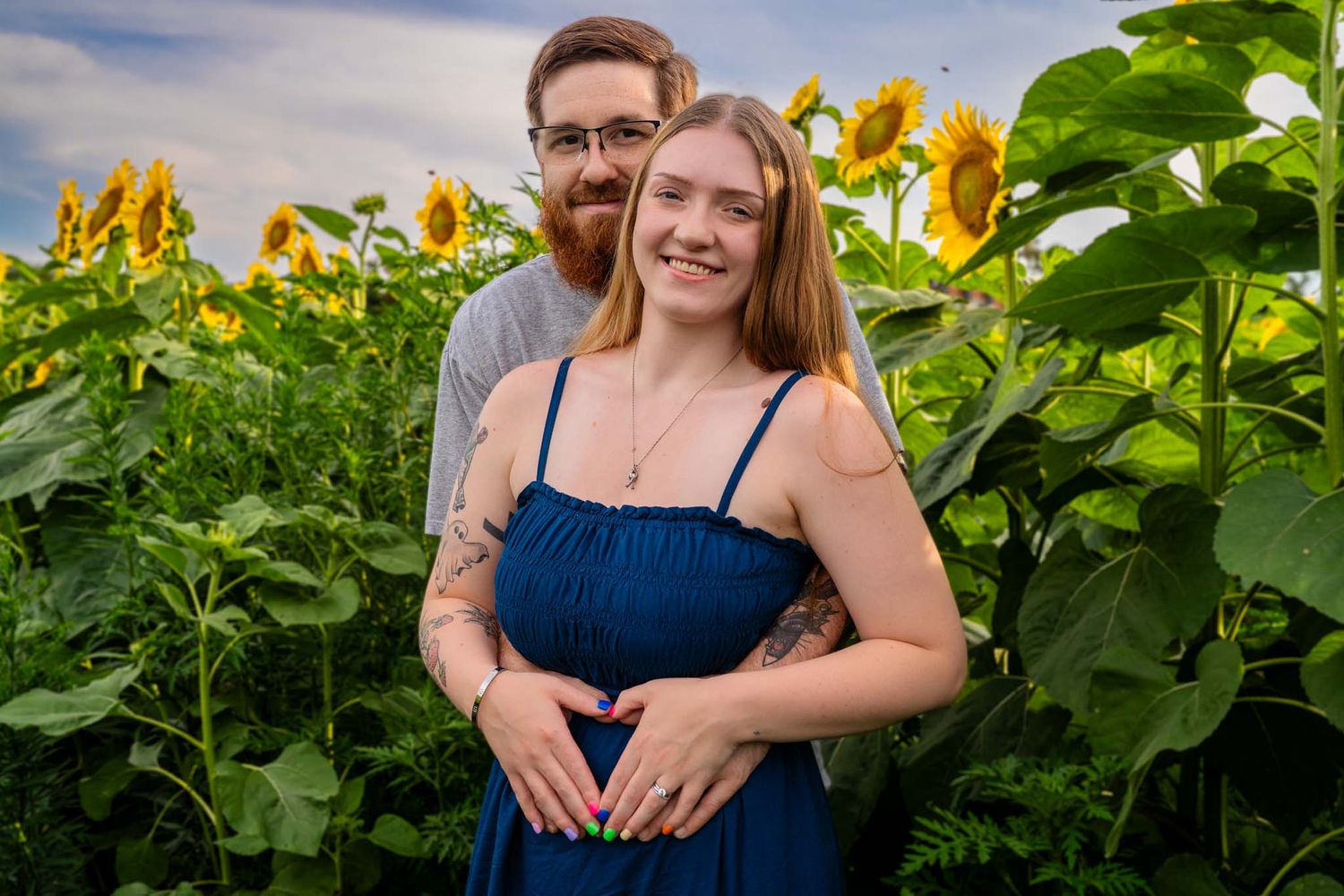 Smiling couple standing in a sunflower field making a heart shape over a baby bump.