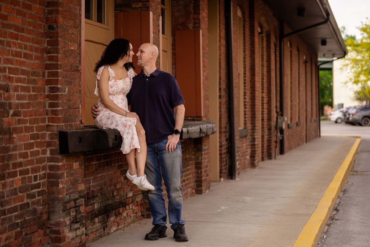 A couple shares a romantic moment outside a rustic brick building, with the woman sitting on a ledge and the man standing close.