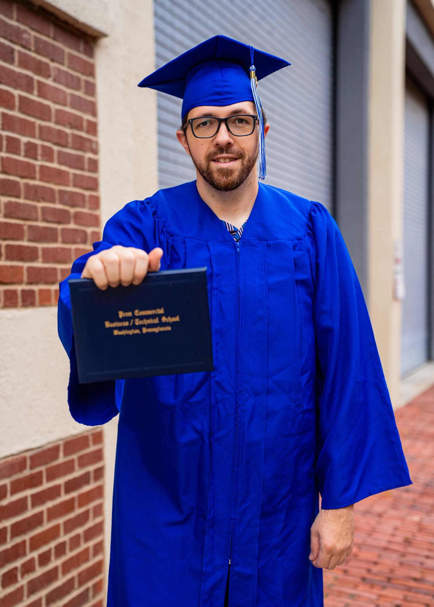Male graduate in blue cap and gown holding diploma outdoors near brick wall.