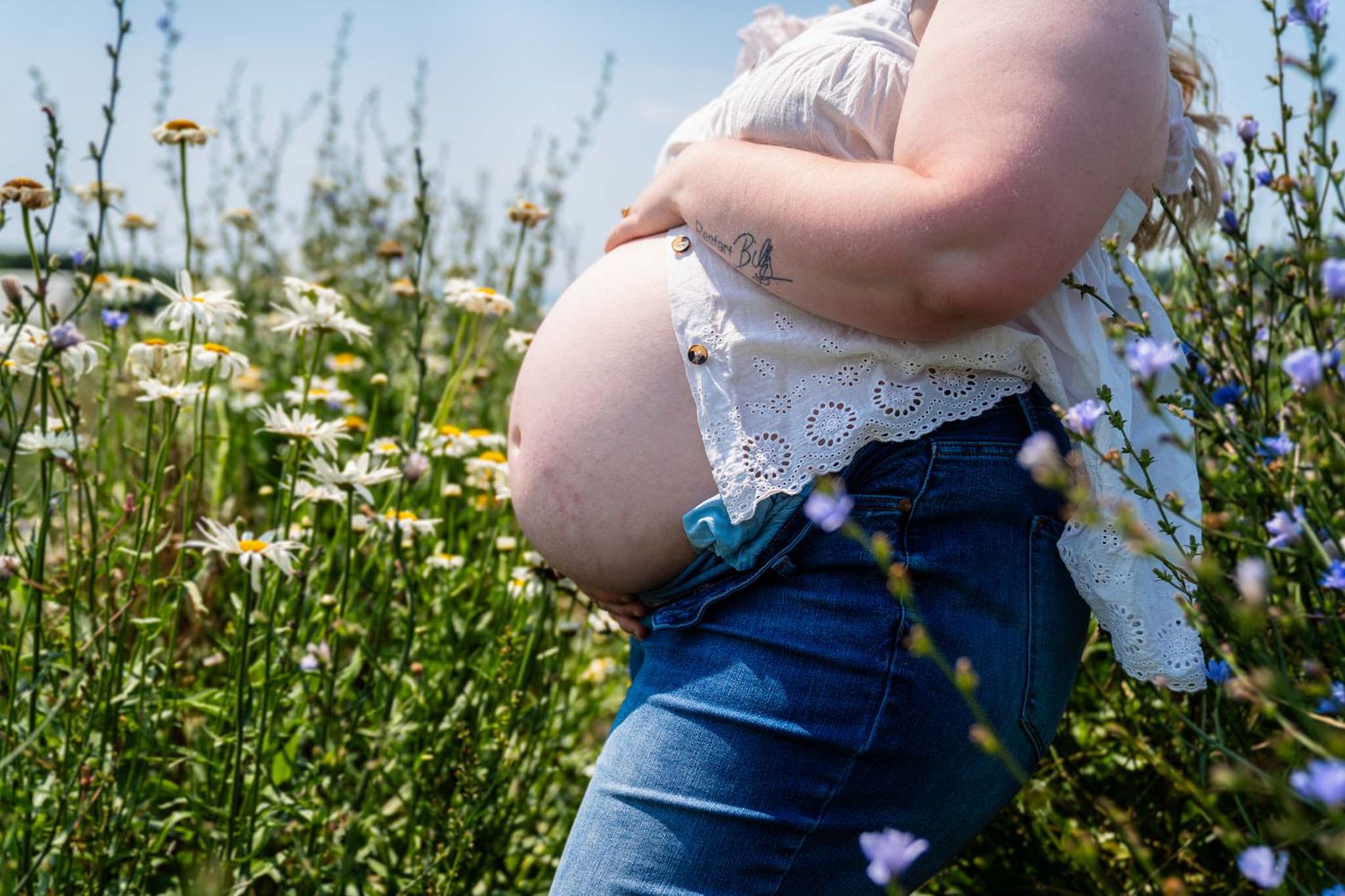 Pregnant woman in white top and jeans standing in a wildflower field with daisies on a sunny day.
