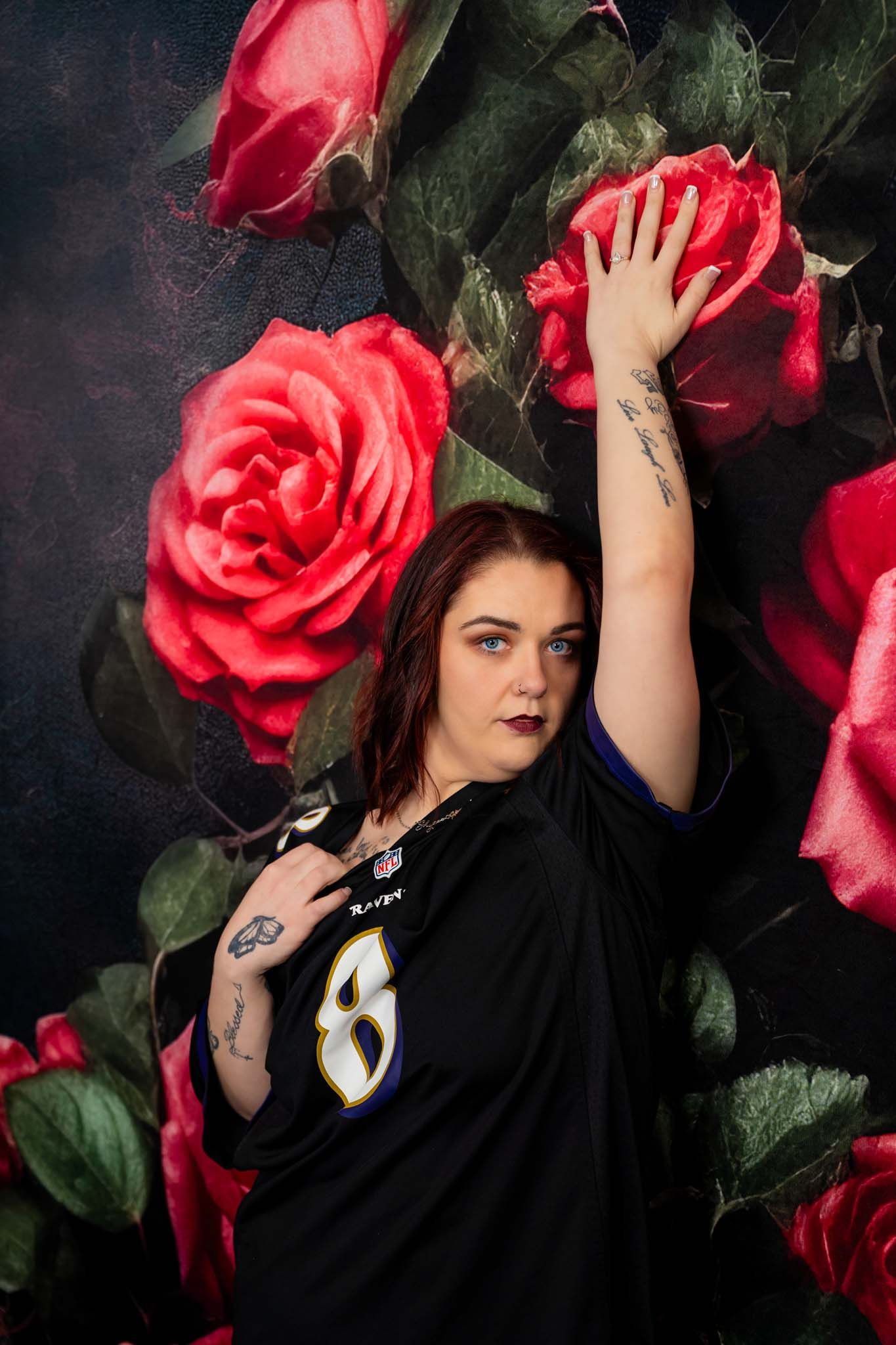 Woman in Ravens jersey poses against floral rose backdrop with arm raised, showcasing tattoos.
