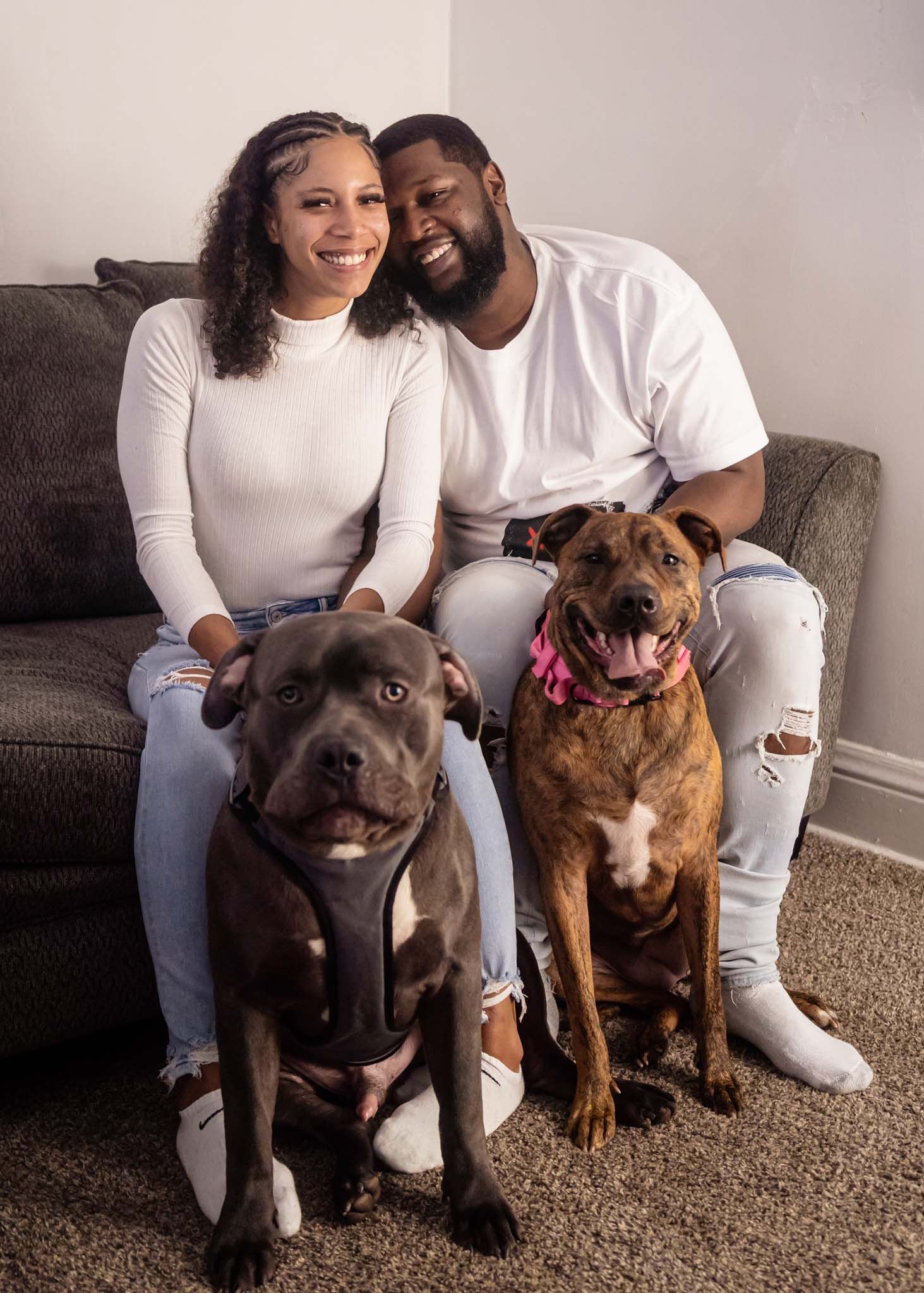 Couple sitting on couch smiling with two pit bull dogs posed in front on carpeted floor.