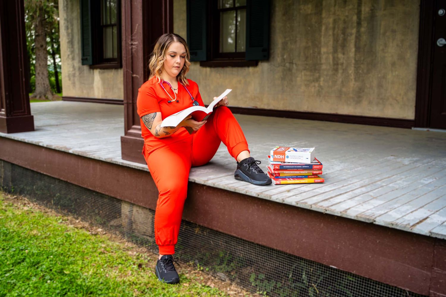 Woman in red jumpsuit reading a book on a porch steps outdoors.