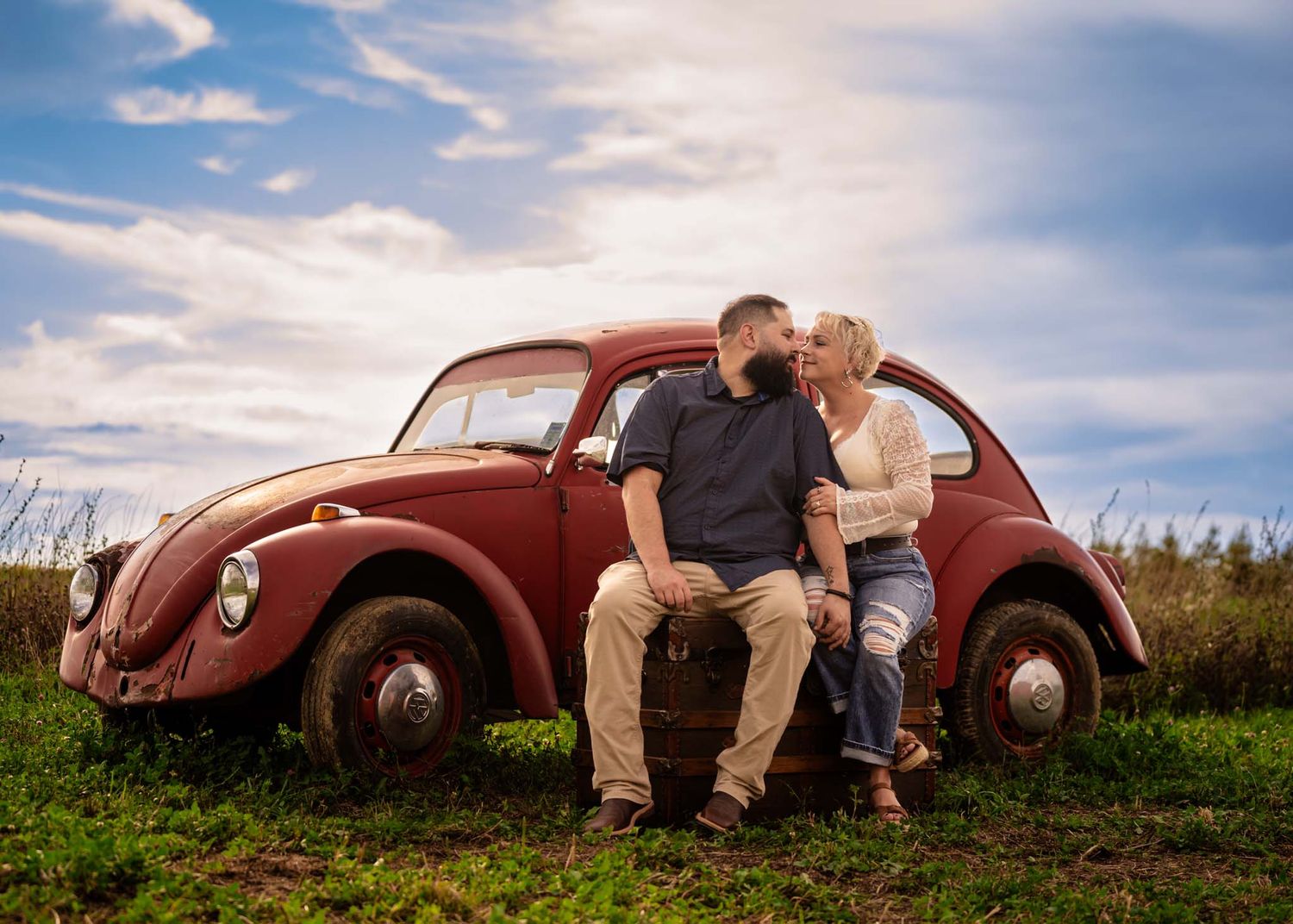 Couple sharing a romantic moment leaning against a vintage red VW Beetle in a green field under a cloudy sky.